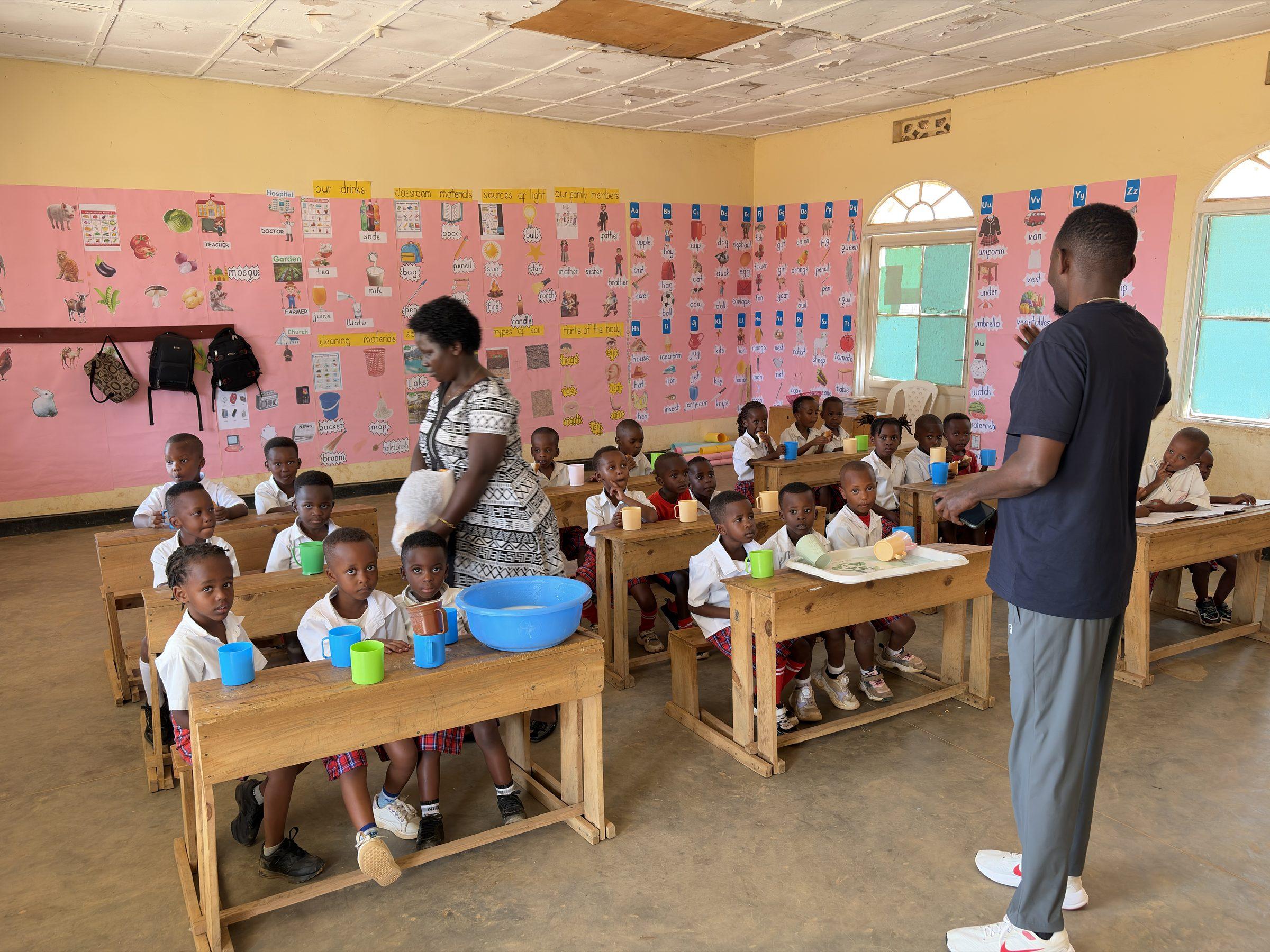 Nursery children with plastic cups at low desks, two staff members serving