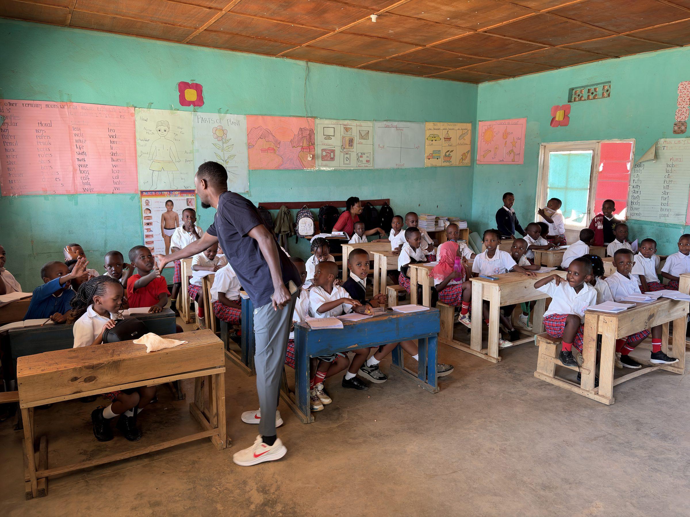 A teacher standing among pupils at wooden desks in a green-walled classroom