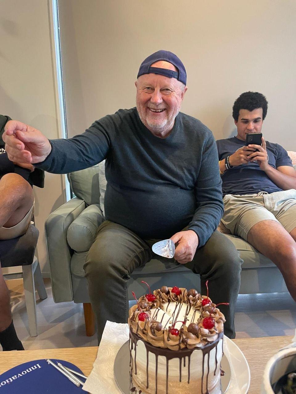 An older man in a cap, seated and smiling, with a cake on the table in front of him