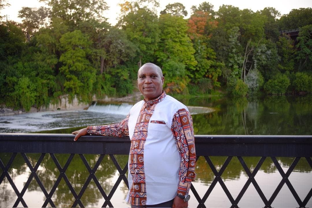 A man in a patterned shirt standing at a railing above a river, trees behind him