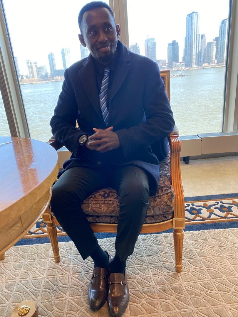 Eddy Kalisa Nyarwaya seated in an armchair in a dark suit and striped tie, a city skyline across the river behind him
