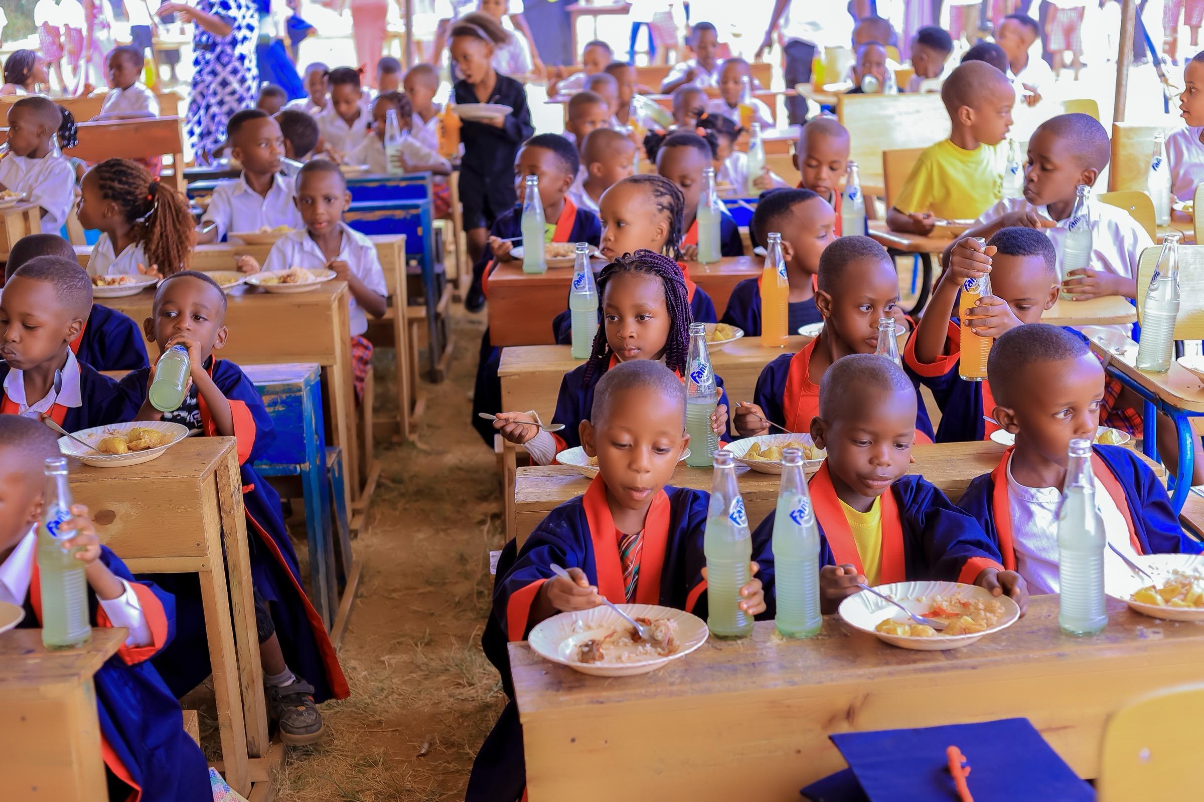 Rows of young children eating lunch at wooden desks under a tent