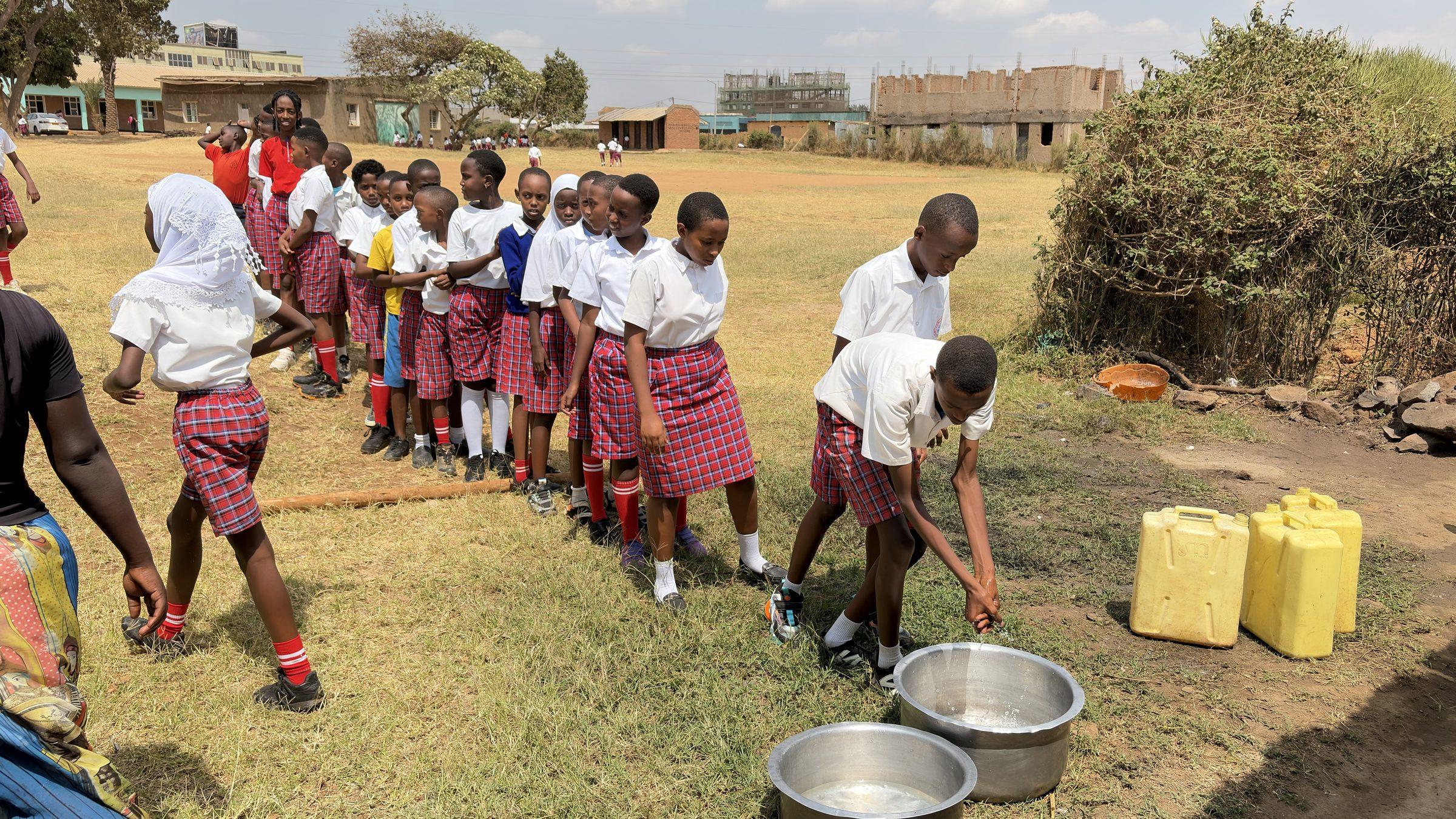 Pupils in a line outside with plastic cups, food pots and yellow jerrycans on the grass