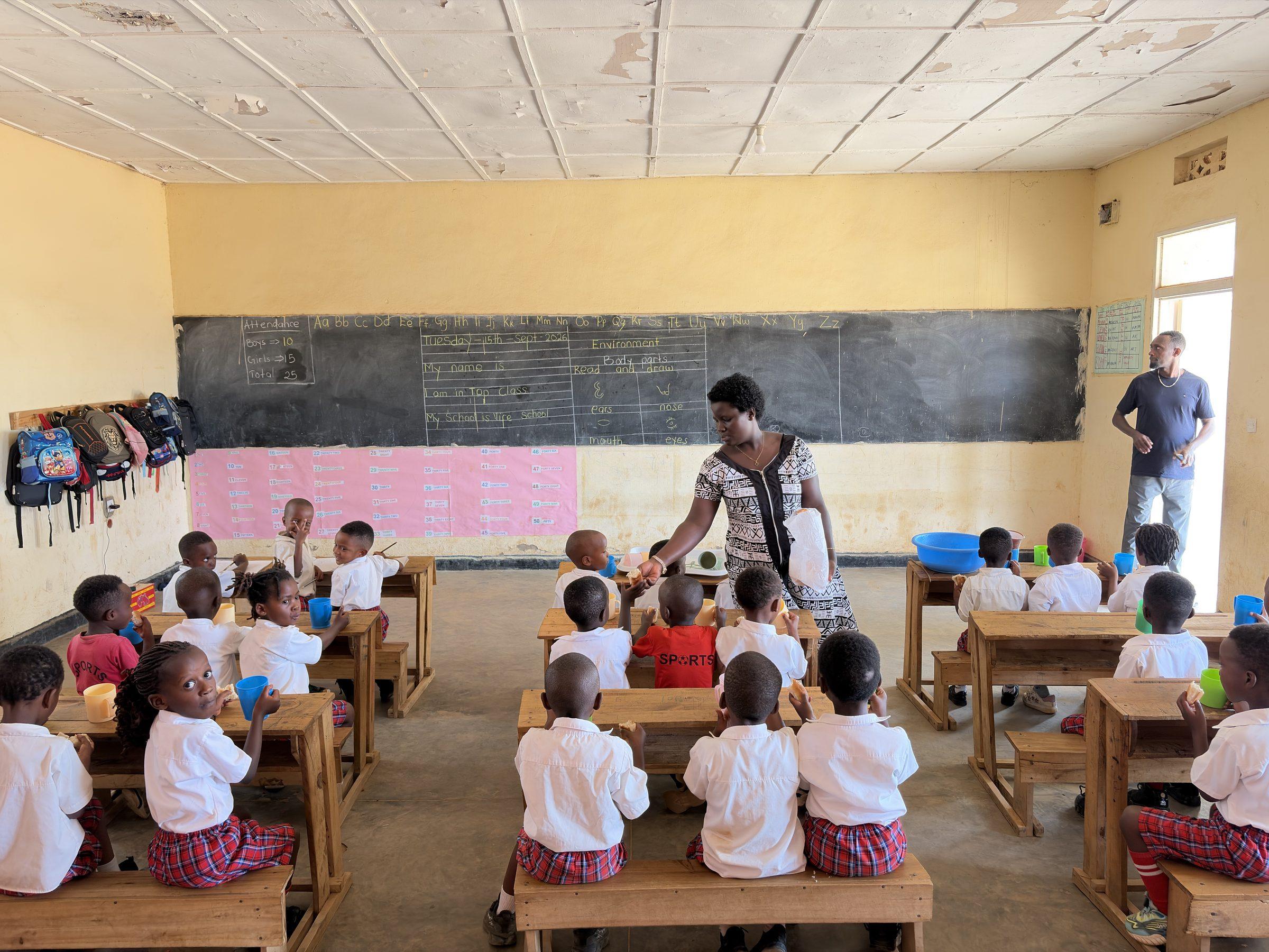 Young children at low desks facing a chalkboard, a teacher among them