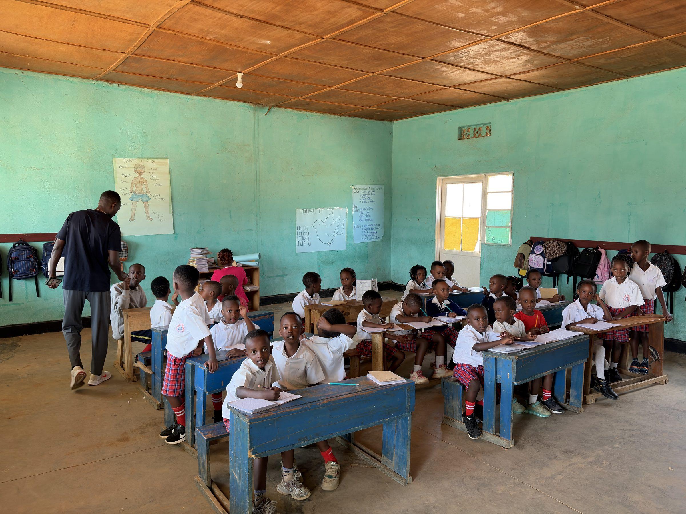A teacher beside the wall charts while pupils work at their desks