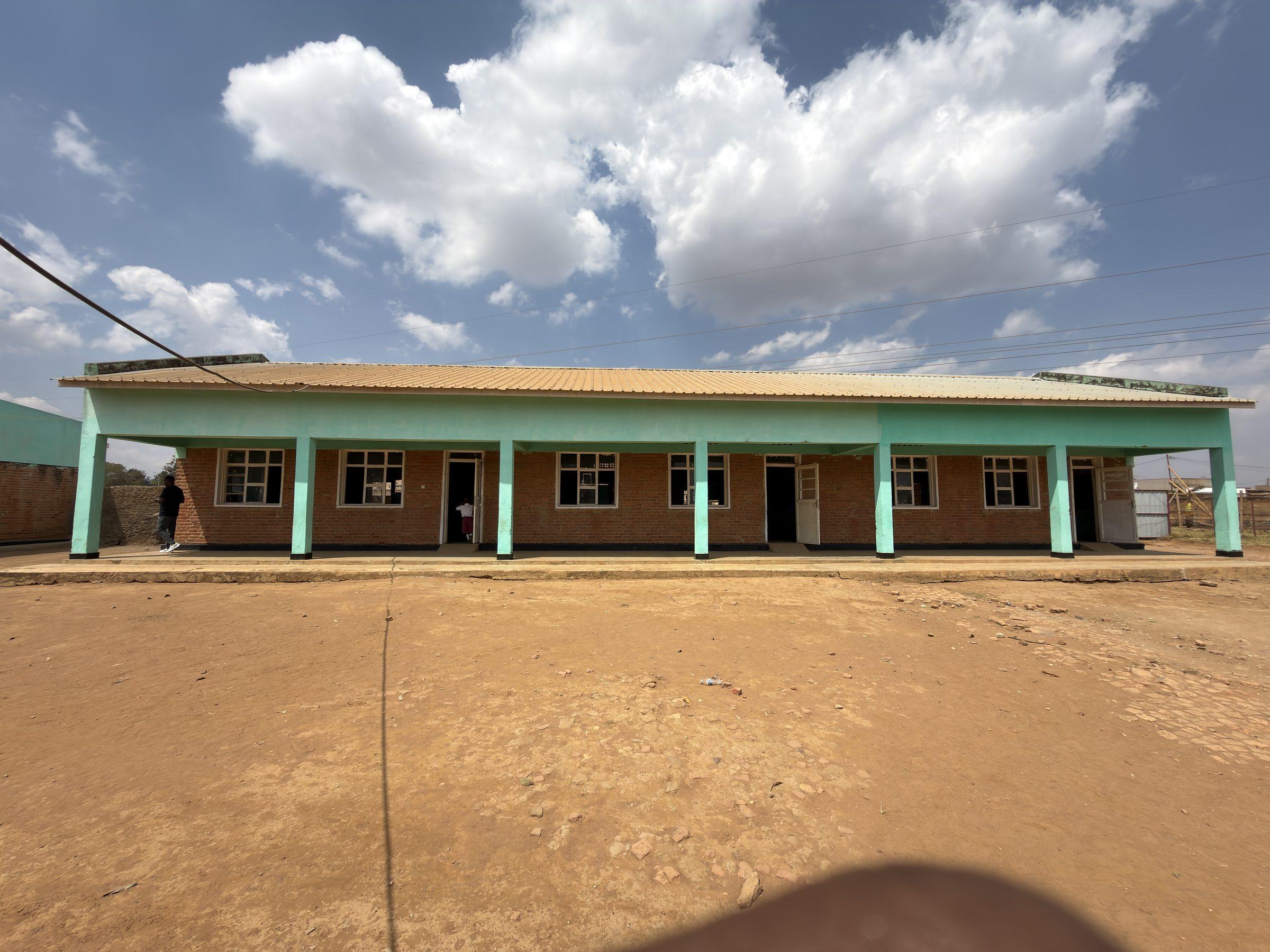 A long single-storey green school building under a wide sky, with an empty earth yard in front